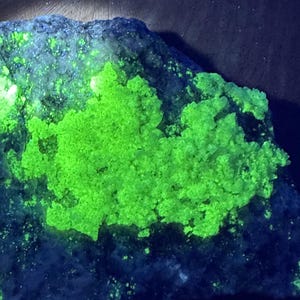Op de afbeelding: Een close-up van een donkerblauwe rots met een felgroene mineraalafzetting. Het mineraal gloeit onder UV-licht.