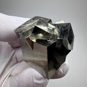 Könnte beinhalten: Eine Ansammlung von metallischen Pyritkristallen mit scharfen, kubischen geometrischen Formen und einer reflektierenden, messinggoldenen Oberfläche. Das Mineralexemplar zeigt ausgeprägte, sich überschneidende quadratische Flächen und glatte Kanten.