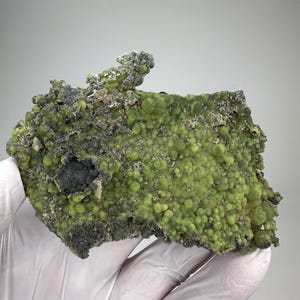 May include: A close-up of a mineral specimen featuring a vibrant, lime-green surface with a bubbly texture. The mineral is set against a neutral background, showcasing its unique, organic form. Dark gray patches add contrast to the overall composition.