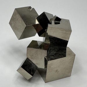 May include: A cluster of metallic pyrite crystals featuring multiple interconnected cubic formations with reflective, brassy surfaces. The mineral specimen displays sharp, geometric edges and smooth, flat faces that catch the light, creating a complex, three-dimensional structure with varying shades of silver and gold.