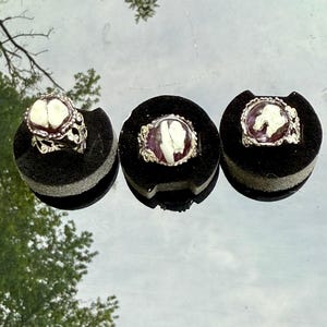May include: Three silver rings with white and purple resin insets. The rings are displayed on black velvet pads.