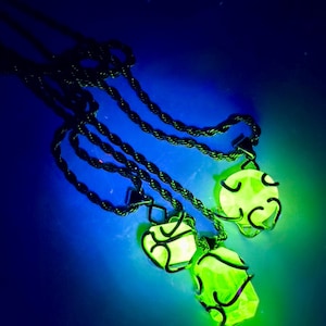 May include: Three glowing green crystal pendants on dark green rope chains. Each crystal is encased in a dark metal wire frame. The background is a gradient of blue and green.