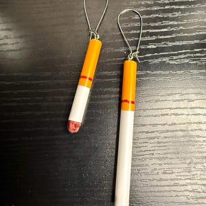 Op de afbeelding: Een paar novelty oorbellen ontworpen om op sigaretten te lijken. Elke oorbel heeft een witte cilindrische behuizing met een oranje punt en een rode band, bevestigd aan een zilverkleurige oorhaak. De uiteinden van de oorbellen zijn gekleurd om op een aangestoken sigaret te lijken.