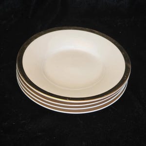 May include: A stack of four round, off-white ceramic bowls featuring a wide, metallic gold rim along the top edge. The bowls have a smooth, shallow interior basin and a clean, simple design, presented as a set of matching dinnerware pieces.