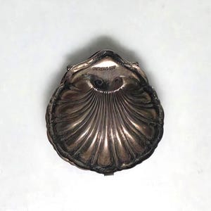 May include: A metallic, shell-shaped dish with a polished, dark silver-toned finish and radiating fluted ridges. The interior surface features the stamped text 925 STERLING near the top edge, indicating the material composition of the object.