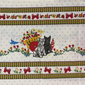 May include: White fabric with a repeating pattern of gray polka dots, floral arrangements, red bows, and a yellow and green plaid border. Two terrier dogs sit near a red wheelbarrow filled with flowers.