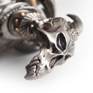 May include: Close-up of a silver-toned metal sculpture featuring a skull with horns. The sculpture has intricate details and a polished finish. The skull's features are clearly defined, with a focus on the horns and facial structure. The overall design suggests a gothic or fantasy theme.