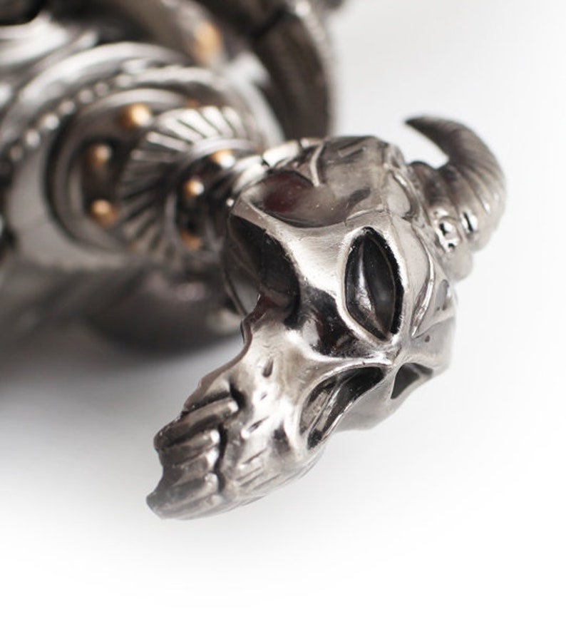 May include: Close-up of a silver-toned metal sculpture featuring a skull with horns. The sculpture has intricate details and a polished finish. The skull's features are clearly defined, with a focus on the horns and facial structure. The overall design suggests a gothic or fantasy theme.