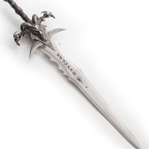 May include: A detailed fantasy sword with a silver blade and intricate hilt. The hilt features a skull design with horns and claw-like elements. The blade has etched inscriptions and a pointed tip, showcasing a medieval or fantasy theme.