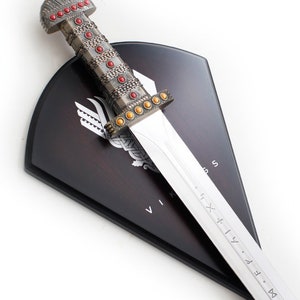 May include: A silver Viking sword with a red and gold hilt mounted on a dark wood shield with a Viking symbol and the word "VIKING" inscribed on it.