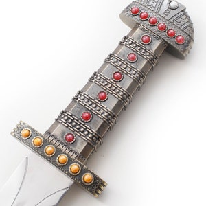 May include: A silver sword with a red and gold pommel. The pommel is decorated with a pattern of circles and squares. The sword has a silver blade and a silver handle.