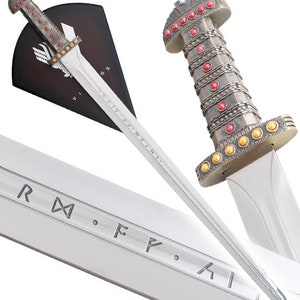 May include: A silver Viking sword with a wooden handle and a red and gold pommel. The sword is displayed on a wooden plaque with the inscription "IX.XI.MM.I.N.I." The sword is a replica of a historical Viking sword.