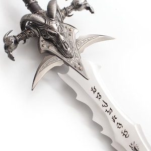 May include: A detailed, silver-colored fantasy sword with a skull-shaped hilt and curved horns. The blade has a series of notches and etched symbols. The hilt features intricate detailing and a leather-wrapped grip.