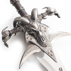 May include: A detailed, silver-colored fantasy sword with a ram skull design on the hilt. The blade is silver with decorative etchings. The handle is wrapped in brown leather and accented with gold-colored studs.