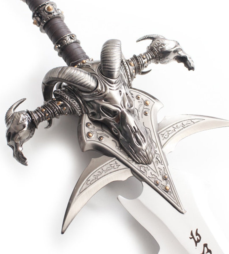 May include: A detailed, silver-colored fantasy sword with a ram skull design on the hilt. The blade is silver with decorative etchings. The handle is wrapped in brown leather and accented with gold-colored studs.
