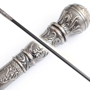 Op de afbeelding: Een zilveren wandelstok met een sierlijke, gedetailleerde handgreep. De handgreep is bolvormig met ingewikkelde bloemmotieven. De stok is zwart en heeft een zilveren punt.