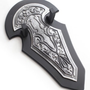 May include: A decorative shield with a black base and a silver-colored, ornate design. The shield features intricate patterns and is mounted on a black stand. The overall aesthetic is reminiscent of medieval or fantasy themes.