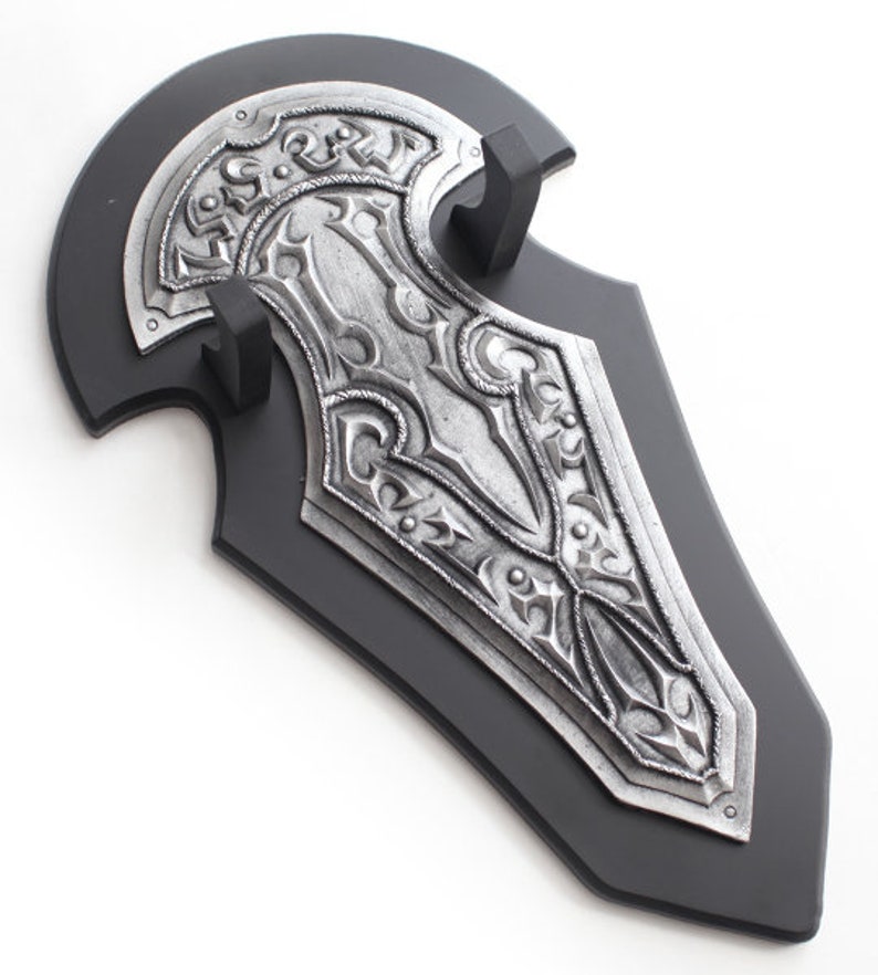 May include: A decorative shield with a black base and a silver-colored, ornate design. The shield features intricate patterns and is mounted on a black stand. The overall aesthetic is reminiscent of medieval or fantasy themes.