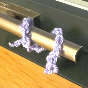 Peut inclure: Gros plan d'un tube métallique doré avec une longueur de fil de laine violet clair enroulée autour. Le fil est noué à deux endroits. Le tube est posé sur un fond sombre et une surface en bois.