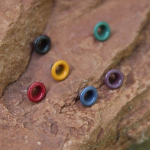 May include: Assortment of colorful metal eyelets in various colors, including red, yellow, blue, and green, arranged on a brown rock surface.