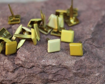 May include: A close-up of a pile of gold-colored metal tacks with square heads. Some of the tacks have a light yellow finish, while others have a darker yellow finish.