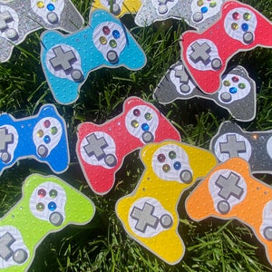 May include: A collection of colorful, decorated game controllers, each with a different design and color scheme. The controllers are made of cardboard and have a textured surface. They are arranged in a semi-circle on a bed of green grass.
