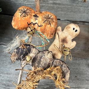 Può includere: Una decorazione di Halloween fatta a mano con una zucca, un fantasma e un pipistrello. Le figure sono fatte di stoffa e hanno un aspetto rustico e vintage. Sono attaccate a un bastone e hanno delle campane appese.