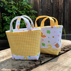 Könnte beinhalten: Zwei kleine gelbe und weiße Gingham-Stofftaschen. Eine Tasche hat ein weißes Stofffutter mit einem Regenbogen, Sternen und Hufeisen darauf gedruckt. Die andere Tasche hat ein weißes Stofffutter mit einem Kleeblatt, Sternen und einem Regenbogen darauf gedruckt.