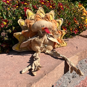 May include: A stuffed fabric turkey figurine featuring a layered fan-shaped tail in shades of yellow and orange, a tan body with burlap accents, a small orange beak, a red wattle, and two long, dangling fabric legs with knotted ends.