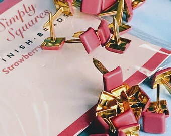 May include: A close-up of a package of pink and gold metal corner brackets. The package is labeled "Simply Squares" and "Finish. Brads. Strawberry".