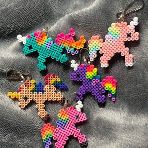 Op de afbeelding: Vijf kleurrijke regenboog-eenhoorn sleutelhangers gemaakt van perler kralen. Elke sleutelhanger heeft een eenhoorn in een andere kleur met een regenboog manen en staart.