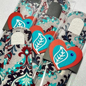 May include: Three gray and white fabric nail files with a paisley pattern. Each file is wrapped in clear plastic and has a red heart with a turquoise leaf and a blue rhinestone on top.
