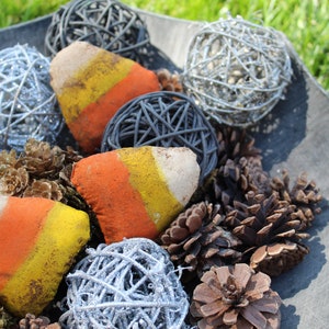 May include: A gray bowl filled with decorative items, including pinecones, white and gray woven balls, and three painted rocks shaped like candy corn.