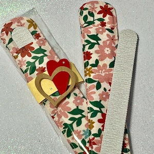 May include: A set of two nail care items, a nail file and a nail buffer, wrapped in a floral fabric with a heart-shaped embellishment. The fabric is pink, white, and green with a floral pattern.
