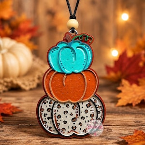 May include: A decorative hanging ornament featuring three stacked pumpkins. The top pumpkin is turquoise, the middle is orange, and the bottom is white with a black leopard print pattern. It hangs from a black cord with a single wooden bead.