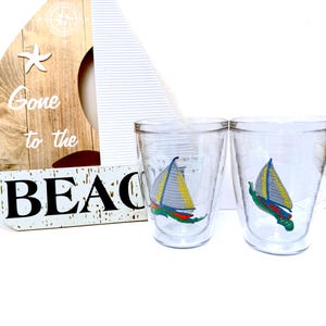 May include: Two clear double-walled tumblers featuring a colorful sailboat graphic with yellow sails and a red and green hull. Behind the tumblers is a wooden sailboat-shaped sign with the text Gone to the BEACH and a white starfish icon.