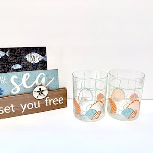 May include: Two clear drinking glasses with a seashell design in orange and teal. The glasses have a grid pattern and are next to a wooden sign that says "Let the sea set you free" with fish and a starfish.