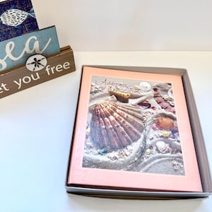 May include: A rectangular address and appointment book with a pink border and a cover image of seashells on sand. Beside it sits a wooden sign with the text "the sea set you free" and a small white sand dollar accent piece attached to the front.