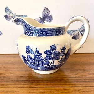 Puede incluir: Una jarra de crema de cerámica blanca con un patrón de sauce azul y blanco. La jarra tiene un cuerpo redondeado y un asa.