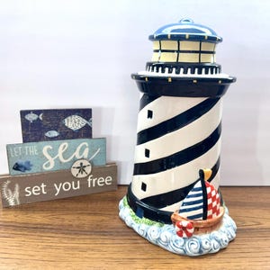 Puede incluir: Un faro de cerámica con rayas blancas y negras, una parte superior azul y un diseño de velero. El faro se asienta sobre una base de olas azules. También hay letreros de madera con el texto "Let the sea set you free".