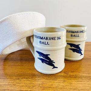 Puede incluir: Dos tazas de cerámica blanquecina con ilustraciones de delfines azules y el texto "SUBMARINE 96 BALL". Un sombrero blanco texturizado se apoya en las tazas. Las tazas están sobre una superficie de madera.