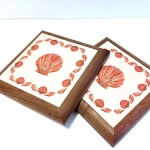 Puede incluir: Dos posavasos cuadrados con fondo blanco, con una gran concha de color coral en el centro y conchas más pequeñas alrededor del borde. Los posavasos tienen un marco de madera marrón oscuro.