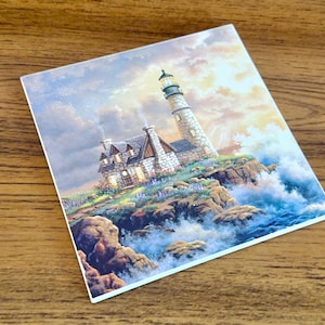 May include: Square coaster featuring a lighthouse scene. The image depicts a tall, white lighthouse with a black top, a house, and crashing waves. The artwork is set against a cloudy sky with a warm, golden light.