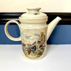 May include: Cream-colored ceramic teapot with a lid and a curved handle. The teapot features a detailed illustration of a seaside scene with boats, buildings, and people. The spout is long and slender, and the lid has a small knob.