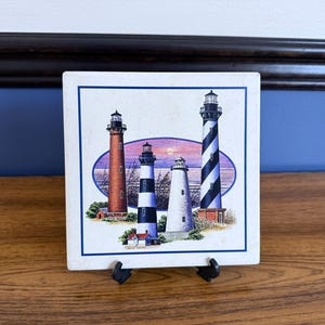 May include: Square ceramic tile featuring four lighthouse illustrations. The lighthouses are depicted in various colours and designs, set against a coastal landscape with a sunset. The tile is displayed on a black easel.