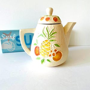 May include: A white ceramic teapot with a lid, decorated with a pineapple, oranges, and green leaves. The teapot has a slightly rounded shape and a spout on the side.