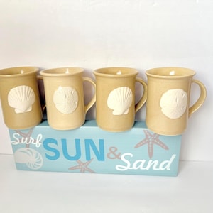 May include: Four beige ceramic mugs with seashell designs are displayed on a blue wooden block that reads "Surf Sun & Sand". The mugs have a handle and a raised seashell design. The block has starfish and seashell graphics.