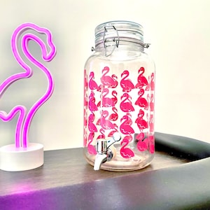 May include: A clear glass beverage dispenser with a silver spigot and lid, decorated with pink flamingo patterns. A pink neon flamingo light is to the left. The dispenser is on a dark surface.