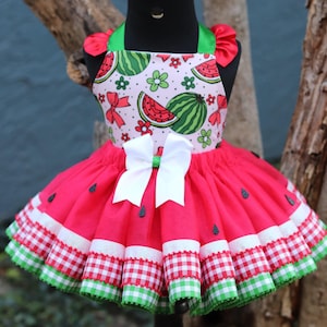 May include: A red and green watermelon-themed tutu dress with a white bow. The dress has a pink bodice with watermelon print and a red tulle skirt with green and white gingham ribbon trim.