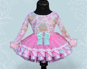 May include: A pink and white long-sleeved dress with a gingerbread house and candy cane pattern. The dress features a pink tulle skirt with candy cane ribbon trim and a light blue bow.